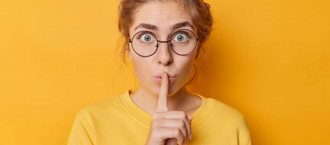 Stunned emotional beautiful woman hushing with index finger shares secret makes taboo gesture stares through round spectacles wears casual jumper isolated over yellow background. Shh be quiet